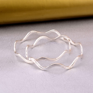 May include: Two silver-colored bangles featuring a continuous wavy, undulating design. The bracelets have a smooth, reflective metallic finish and a circular shape, presented as a pair of matching accessories with a consistent, flowing pattern.