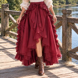 Pode incluir: Uma saia assimétrica vermelho-escura com bainha com babados e fecho de laço na cintura. A saia tem um design em camadas com painéis de tecido em cascata que criam uma silhueta volumosa, combinada com botas de couro castanho.