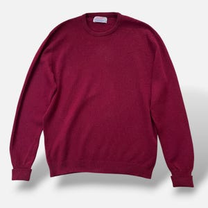 Könnte beinhalten: Ein langärmeliger Pullover mit Rundhalsausschnitt in einem tiefen Burgunderrot. Das Kleidungsstück verfügt über einen gerippten Strickkragen, Bündchen und einen Saum. Ein kleines weißes rechteckiges Markenetikett ist am inneren Halsausschnitt eingenäht.