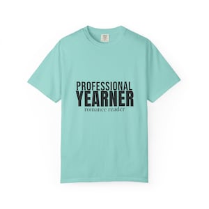 Può includere: Una t-shirt a maniche corte color turchese chiaro con scollo rotondo. Al centro del petto è presente una scritta nera che recita PROFESSIONAL YEARNER in un carattere sans-serif in grassetto, con romance reader in un carattere serif più piccolo.