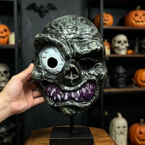 May include: A full-face Halloween mask featuring a textured dark green and black finish. The mask has one large, bloodshot white eye, a hollow eye socket, and a wide, grinning mouth filled with prominent, glossy purple teeth and gums.