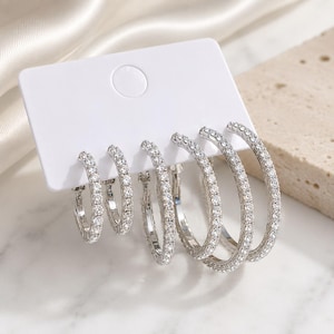 May include: A set of three pairs of silver-toned hoop earrings displayed on a white rectangular card. Each hoop is circular and features a continuous row of small, clear, sparkling stones embedded along the entire outer surface of the metal.