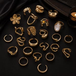 Può includere: Una collezione di ventidue anelli in metallo color oro dal design vario, inclusi fasce aperte, superfici testurizzate, una forma a fiore, una pietra perlata e un anello con gemme multicolori, disposti su una superficie scura.