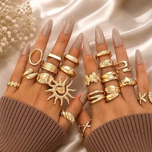 Pode incluir: Uma coleção de anéis dourados usados nos dedos, com vários designs, incluindo uma forma de sol, padrões florais, bandas lisas, texturas torcidas e formas de coração. Os anéis são feitos de metal polido e são usados em vários dedos.