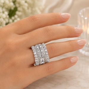May include: A wide silver-toned band ring featuring three rows of sparkling clear stones. The center row consists of rectangular-cut stones, while the two outer rows are composed of smaller, round-cut stones, creating a multi-faceted, brilliant appearance.