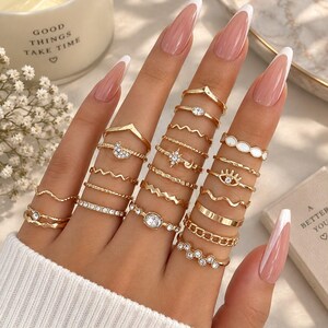 May include: A collection of gold-toned rings worn on four fingers, featuring various designs including a crescent moon with crystals, a starburst, an eye motif, chain links, wavy bands, and clear stones set in metal bands of different widths and textures.