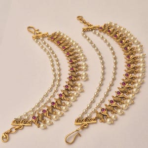 May include: A pair of gold-toned ear chains featuring multiple strands of white faux pearls and ornate metal accents with embedded magenta stones. Each piece includes a hook at the top and bottom for attachment, showcasing a curved, layered design.