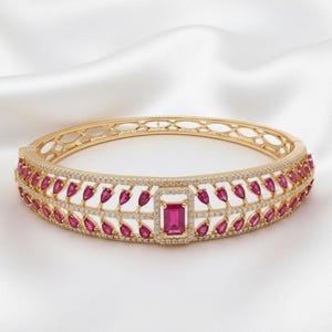 Puede incluir: Un brazalete rígido de oro con bisagra que presenta una piedra preciosa roja central de corte esmeralda rodeada de pequeñas piedras blancas. La banda muestra un patrón simétrico de hojas con piedras rojas en forma de pera.