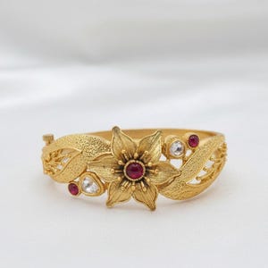 Puede incluir: Un brazalete rígido articulado en tono dorado con un motivo floral central que presenta una piedra roja oscura en el centro. La superficie metálica texturizada está decorada con pequeñas piedras blancas transparentes y piedras rojas redondas.