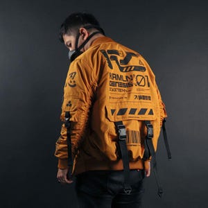 Puede incluir: Una chaqueta bomber de color amarillo mostaza con estampados gráficos en negro, incluyendo el texto "KRMLN GENESIS SYSTEM", un código de barras y caracteres japoneses. La chaqueta incluye correas con hebillas negras en la espalda y mangas.