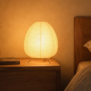 May include: A round, off-white paper table lamp with a ribbed texture and thin metal legs, emitting a warm, soft glow. The lamp sits on a wooden nightstand next to a small, dark ceramic vase containing dried stems and a closed book.