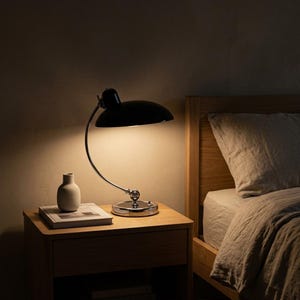 May include: A black desk lamp with a curved chrome arm and a circular base, emitting a warm glow. The lamp sits on a wooden nightstand next to a small white vase placed on top of a book, positioned beside a bed with a light-coloured linen pillow.