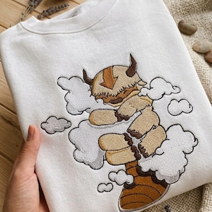 May include: A white crewneck sweatshirt featuring a central embroidered design of a tan, furry, horned creature with a brown arrow marking on its head, depicted flying amidst white clouds with a brown tail visible at the bottom of the graphic.