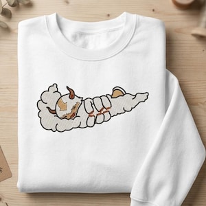 May include: A white crewneck sweatshirt featuring a central embroidered graphic of a stylized, fluffy, six-legged creature with brown horns and a brown arrow marking on its head, depicted in a flying pose surrounded by soft, cloud-like shapes.