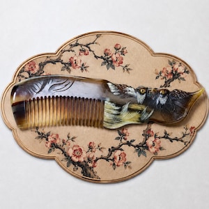 Puede incluir: Un peine de cuerno marrón oscuro con un intrincado diseño de búho tallado en el mango, con plumas detalladas y ojos amarillos. El peine se muestra sobre una placa ovalada beige decorada con un patrón pintado de ramas marrones y flores rosas.