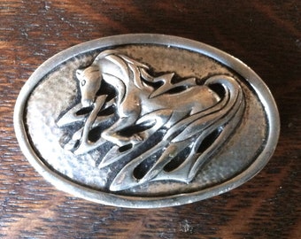 May include: Oval-shaped silver-toned belt buckle featuring a detailed relief of a horse in motion. The horse design is set against a textured background, with stylized flames incorporated into the design. The buckle is a Western-style accessory.