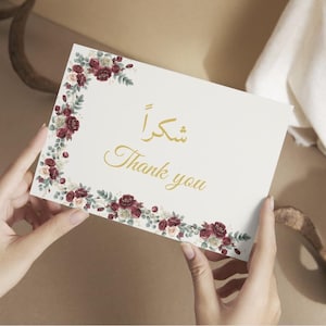 May include: A rectangular white card featuring a floral border of deep red and cream roses with green leaves. The card displays the text Thank you in gold script, accompanied by the Arabic word for thank you in matching gold lettering.
