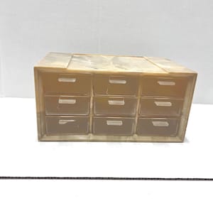 May include: A rectangular storage organizer featuring a marbled tan and beige exterior finish. The unit contains nine individual drawers arranged in three columns and three rows, each equipped with a small, clear, horizontal pull handle.