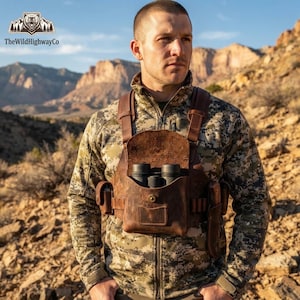 Peut inclure: Un harnais de poitrine en cuir marron doté d'une pochette centrale contenant des jumelles noires, des emplacements pour accessoires sur les côtés et des bretelles réglables, porté sur une veste zippée à manches longues à motif camouflage.