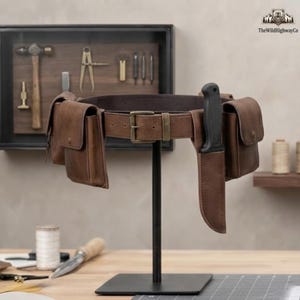 Peut inclure: Une ceinture utilitaire en cuir marron dotée d'une boucle en laiton, de deux poches latérales à boutons-pression et d'un étui en cuir contenant un couteau à manche noir. La ceinture est présentée sur un support en métal noir.