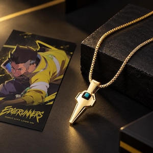May include: A gold-toned pendant necklace featuring a geometric, tapered design with a small, square turquoise-coloured inlay. The pendant hangs from a matching gold-toned box chain. Beside it lies a rectangular card with an anime-style illustration.
