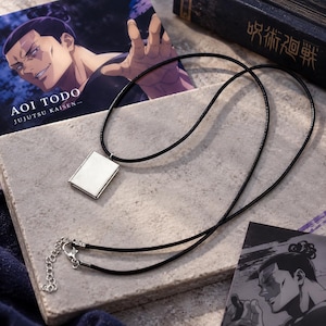 Può includere: Un ciondolo rettangolare a forma di libro color argento sospeso su una sottile collana con cordino nero. Il ciondolo presenta una superficie metallica liscia con un dettaglio a cerniera sul lato. La collana include una chiusura a moschettone in metallo argentato e una catenella di estensione regolabile.