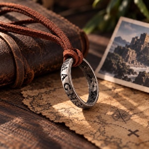 Puede incluir: Un colgante de anillo de metal en tono plateado con un acabado texturizado y desgastado, grabado con letras, suspendido de un cordón de cuero marrón. El anillo presenta caracteres visibles, incluidas las letras M, A, G y C en su superficie.