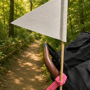 May include: A white triangular felt flag attached to a wooden pole, secured to a stroller handle with a bright pink silicone strap. The stroller features a brown leather-textured handle and black fabric, positioned outdoors on a path.