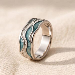 May include: A silver-toned ring featuring a band with a wavy, recessed design. The surface of the ring is accented with a smooth, teal-coloured enamel inlay that follows the undulating pattern, contrasted by a textured, shimmering silver section.
