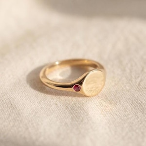 Pode incluir: Um anel de sinete dourado com um topo circular liso e plano, e uma única pequena pedra preciosa redonda de cor rosa vibrante cravada na banda logo ao lado da face circular, apresentando um acabamento metálico polido e design simples.