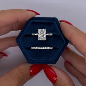 Puede incluir: Un anillo de compromiso con diamante de corte radiante y banda pavé, junto a una alianza de boda pavé a juego, presentados en un estuche hexagonal de terciopelo azul oscuro con dos ranuras horizontales para los anillos.