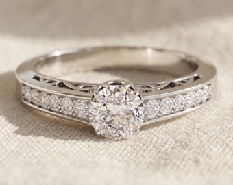 May include: A silver-toned ring featuring a large, round, faceted center stone held in a prong setting. The band is accented with a channel of smaller, round, sparkling stones on both sides and displays intricate openwork detailing along the sides.