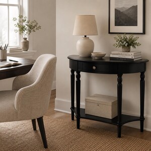 May include: A black semi-circular console table with a single drawer featuring a small round knob. The table has four turned legs and a lower shelf holding a rectangular beige storage box. The top surface holds a lamp and a small bowl.