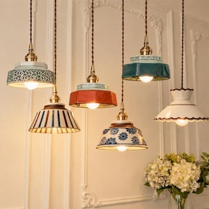 Könnte beinhalten: Fünf hängende Pendelleuchten mit Keramikschirmen in verschiedenen Farben und Mustern, darunter blau-weiße Blumenmuster, einfarbiges Orange, Petrol und Weiß mit gewelltem Rand. Jede Lampe hat eine messingfarbene Halterung und ein gedrehtes braunes Kabel.