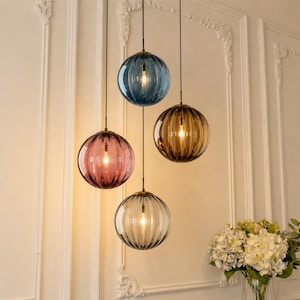 May include: A cluster of four spherical pendant lights with ribbed glass shades in blue, amber, pink, and clear tones. Each globe hangs from a thin black cord at varying heights, containing a single illuminated bulb that casts a warm glow.