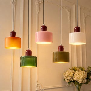 Pode incluir: Cinco candeeiros suspensos cilíndricos pendurados por cabos pretos. Cada lâmpada apresenta um abajur de vidro de cor sólida em laranja, verde, rosa, azeitona e branco, rematado com uma tampa vermelha escura. Emitem uma luz quente.