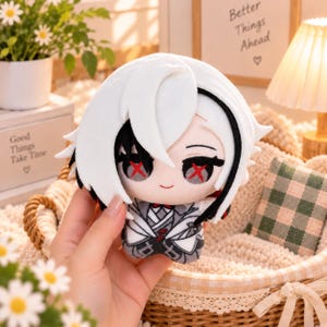May include: A plush doll featuring white hair with black accents, large eyes with red cross-shaped pupils, and a small smile. The doll wears a gray and white patterned outfit with a red bow tie, crafted from soft fabric with detailed embroidery.