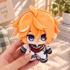 May include: A plush doll featuring bright orange hair, large blue eyes, and a small pink mouth. The doll wears a white and dark gray outfit with a maroon sash, intricate black line details, and a small blue gem accent on the chest area.