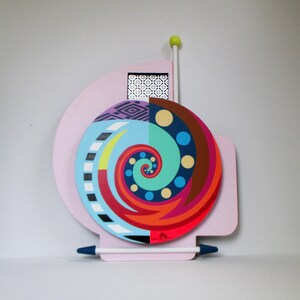 以下が含まれることがあります： カラフルな抽象的な壁の芸術作品で、スパイラルデザインが特徴です。このアートワークは、青、赤、緑、茶色の色合いの円形デザインのピンク色の背景を特徴としています。緑色のボールが付いた白い棒と、白いペン状のオブジェクトも存在します。