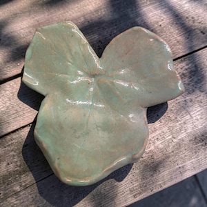 May include: A hand-formed ceramic dish shaped like a three-lobed leaf. The surface features a glossy, light sage green glaze with subtle brown undertones visible along the edges and veins, creating a smooth, reflective finish on the clay form.