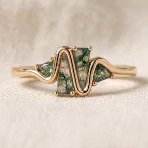 May include: A gold ring featuring a wavy, serpentine band set with four triangular moss agate stones. The stones display translucent white backgrounds with dark green, fern-like inclusions, held securely by small gold prongs along the band.