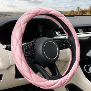 May include: A light pink steering wheel cover featuring a quilted diamond pattern. The cover is installed over a black steering wheel with integrated control buttons and a circular center hub, positioned within the interior of a vehicle.