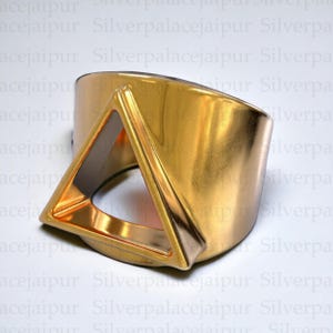 Peut inclure: Un large bracelet manchette doré et poli, doté d'une découpe triangulaire saillante sur le devant. La surface est lisse et réfléchissante, présentant une finition métallique qui capte la lumière sur toute sa structure incurvée et solide.