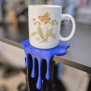 May include: A white ceramic mug featuring a botanical illustration of yellow flowers and the text Primula officinalis, resting on a blue coaster designed to look like a liquid spill dripping over the edge of a flat surface.