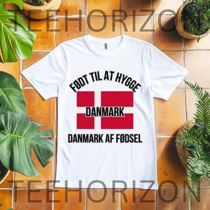 May include: A white short-sleeved t-shirt featuring the text FØDT TIL AT HYGGE at the top, a graphic of the red and white Danish flag with the word DANMARK centred on it, and the text DANMARK AF FØDSEL printed in black lettering below the flag.