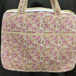 Op de afbeelding: Een gewatteerde laptoptas met een bloemenpatroon in roze, witte en beige tinten. De tas heeft twee handvatten met een geel-wit gestreepte bies, een witte ritssluiting en een horizontaal voorvak met bijpassende gestreepte afwerking.
