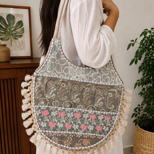 Könnte beinhalten: Eine große, abgerundete Umhängetasche mit einem Patchwork-Design aus grauen, weißen und rosafarbenen Blumen- und Paisleymustern. Die Tasche ist mit kleinen Pailletten, goldfarbenen Perlen und einer Fransenborte aus cremefarbenen Quasten verziert.