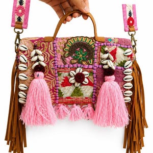 Könnte beinhalten: Eine rechteckige Handtasche aus einem Patchwork aus rosa, roten und grünen bestickten Stoffen. Die Tasche ist mit weißen Kaurimuscheln, zwei großen rosa Quasten und langen braunen Fransen an den Seiten verziert. Sie verfügt über einen Tragegriff und einen abnehmbaren, bestickten rosa Schulterriemen.