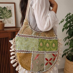 Könnte beinhalten: Eine halbmondförmige Umhängetasche mit einem Patchwork-Design aus verschiedenen bestickten Stoffen in Grün-, Gold- und Rottönen. Die Tasche ist mit weißen Quasten entlang der geschwungenen Unterkante verziert und hat einen breiten, passenden Schulterriemen.