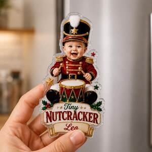 May include: A clear acrylic magnet featuring a smiling baby dressed in a red and gold nutcracker uniform with a tall black hat, playing a drum. The design includes green holly leaves, red berries, and the text Tiny NUTCRACKER and the name Leo.